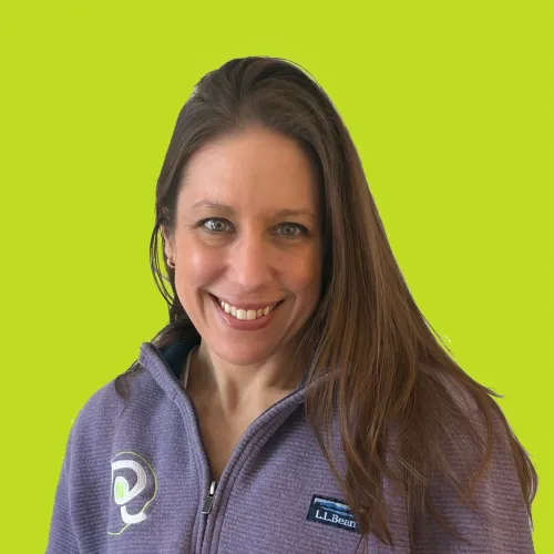Blythe Yost, owner of Rockfield Landscape, smiling while holding a potted green plant. She is wearing a red plaid shirt and a smartwatch, standing against a light gray background.